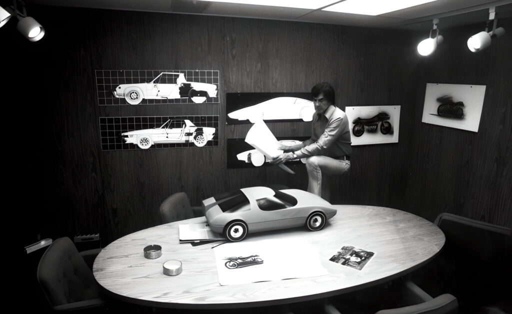John DeLorean, in the early days of DeLorean Motor Company, reviewing a blueprint, standing in front of an automotive clay model, with car schematics and motorcycle renderings on the wall and table.